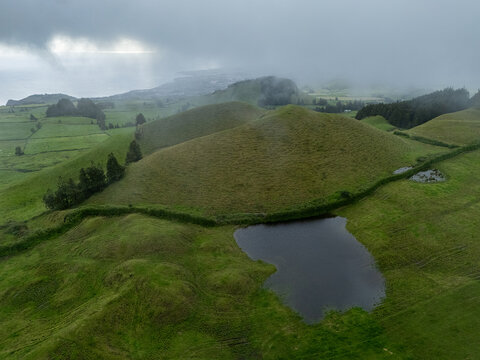 Aerial view of Lagoa Pau Pique surrounded by lush green rolling hills and volcanic landscape under a cloudy sky in Capelas, Azores, Portugal.