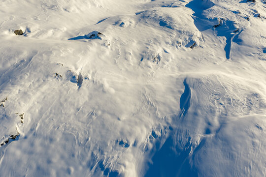 Aerial view of snow-covered mountain terrain with wind-swept patterns, rocky outcrops, and deep shadows in Valldal, More og Romsdal, Norway.