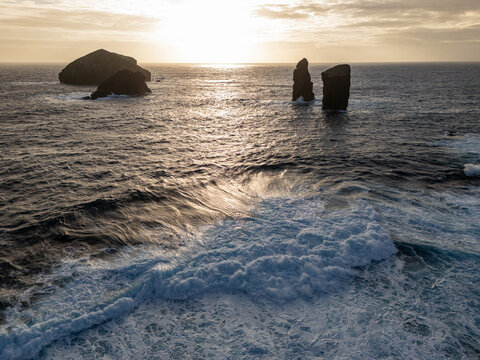 Aerial view of Ilheu dos Mosteiros with waves crashing against the rocky islets during golden hour in Mosteiros, Azores, Portugal.