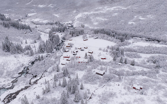 Aerial view of Ostestolen featuring small red cabins nestled in a snow-covered landscape with pine trees and a frozen stream in Valldal, More og Romsdal, Norway.