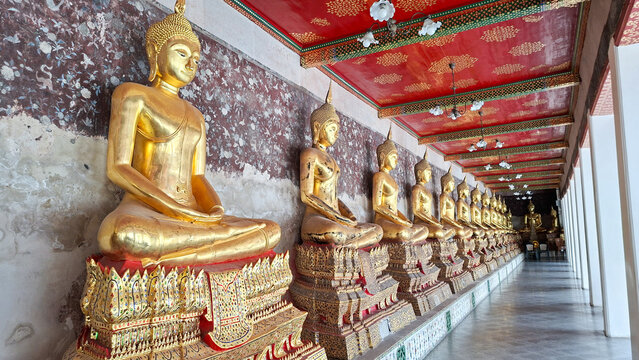 row of golden seated buddha statues in meditation posture along cloister gallery corridor with red ceiling and ancient mural wall at Wat Suthat on August 12, 2026 in Bangkok, Thailand.