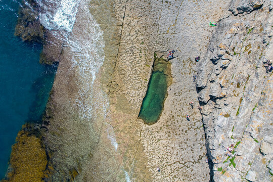 Aerial view of Dancing Ledge, a natural rock pool carved into the limestone cliffs with people swimming and sunbathing on the rocky coast in Swanage, England, United Kingdom.