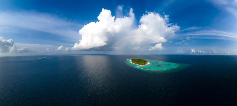 Aerial view of Meedhupparu island surrounded by turquoise lagoon and deep blue ocean under a large white cloud with a faint rainbow in Raa Atoll, Maldives.