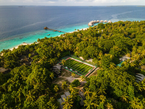 Aerial view of a football pitch nestled among lush palm trees on the tropical island of Meedhupparu, with overwater bungalows and turquoise ocean waters in Raa Atoll, Maldives.