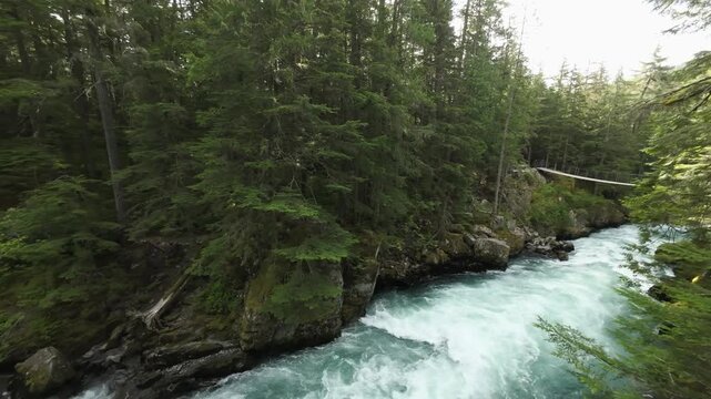 A low aerial camera sweeps out from a green woodland canopy, flying over roaring turquoise rapids to reveal a rustic suspension footbridge over a rocky canyon on a summer day in Whistler.