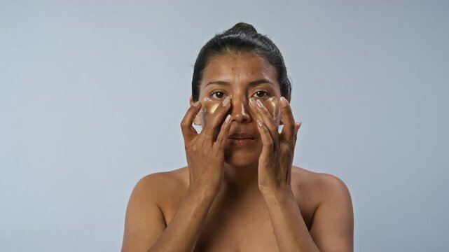 Portrait of Hispanic young woman applying golden hydrogel eye patches under eyes and smiling. Video is perfect for skincare brand advertising and beauty marketing