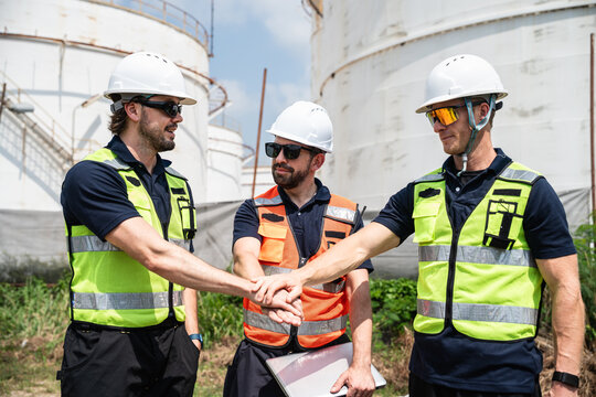Refinery Engineering Team Stacking Hands in a Teamwork Gesture while Wearing Hard Hats and Reflective Safety Vests in Front of Large Petroleum Storage Tanks at an Industrial Facility