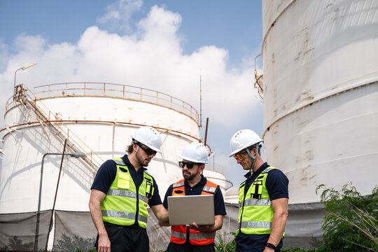 Refinery Engineering Team Reviewing Laptop Data while Wearing Hard Hats and Reflective Safety Vests beside Large Petroleum Storage Tanks at an Outdoor Petrochemical Facility