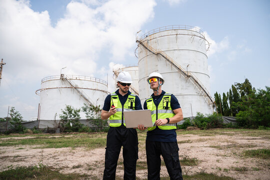 Refinery Engineers Reviewing Operational Data on a Laptop while Wearing Hard Hats and Reflective Safety Vests in Front of Large Petroleum Storage Tanks at an Industrial Facility