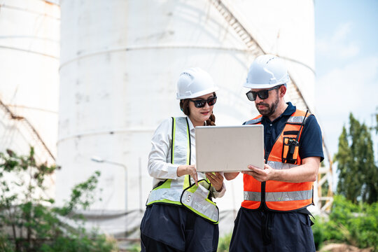 Refinery Engineers Reviewing Operational Data on a Laptop Computer while Wearing Hard Hats and Reflective Safety Vests in Front of Large Petrochemical Storage Tanks at an Industrial Facility