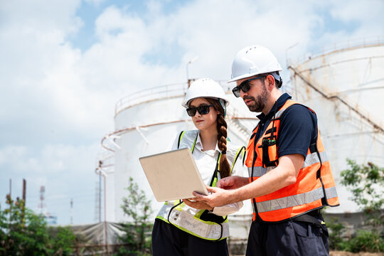 Refinery Engineers Conducting a Field Inspection with a Laptop Computer while Wearing Hard Hats and Reflective Safety Vests beside Petroleum Storage Tanks at an Outdoor Industrial Facility
