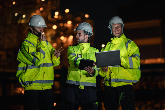 Refinery Operations Team Coordinating Evening Work with a Laptop, Digital Tablet, and Handheld Radio while Wearing Reflective Jackets and Hard Hats at a Petrochemical Plant