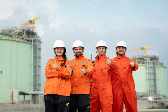 Refinery Engineering Team Standing in Orange Safety Coveralls and White Hard Hats with Thumbs Up in Front of Large Industrial Storage Tanks at a Petrochemical Facility Site