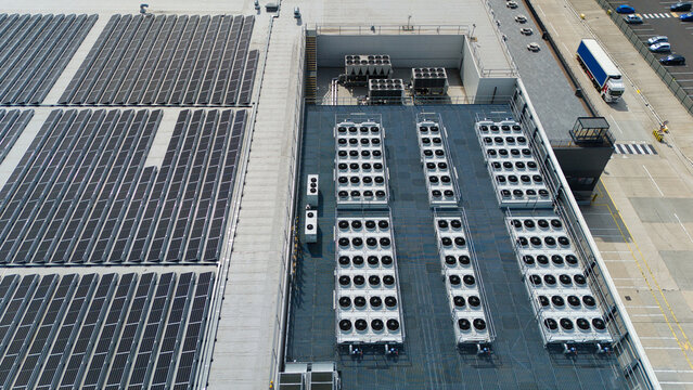Aerial view of ventilation cooling units, heat exchangers, and HVAC fans installed on the roof of an industrial building next to large solar panel arrays in Luton, England, United Kingdom.