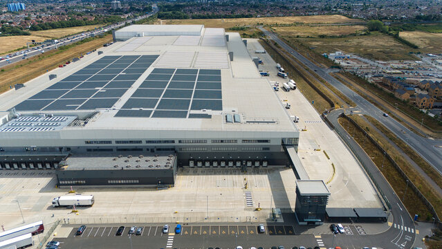 Aerial view of a large supermarket distribution centre featuring extensive rooftop solar panels, numerous loading bays, and a busy parking area in Luton, England, United Kingdom.