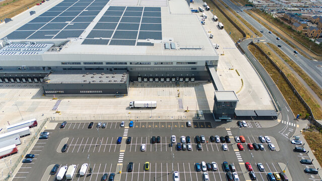 Aerial view of a supermarket distribution centre with a large warehouse, solar panels, loading bays, and a busy car park in Luton, England, United Kingdom.
