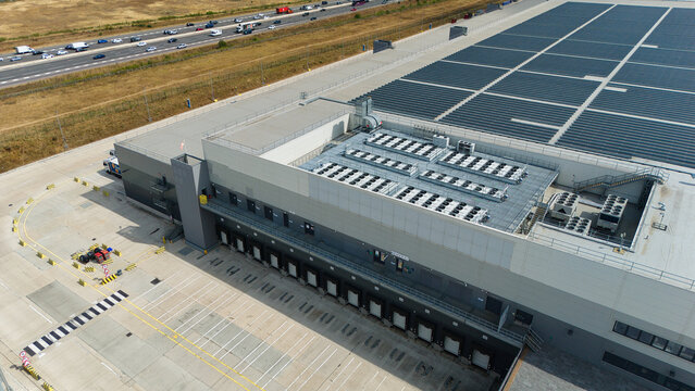 Aerial view of a modern supermarket distribution centre with loading bays, a large flat roof with industrial HVAC units, and adjacent highway traffic in Luton, England, United Kingdom.