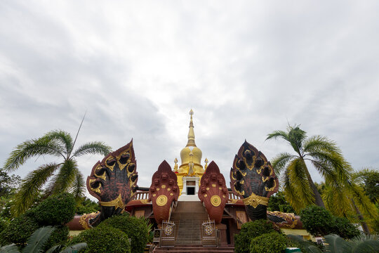 Wide-angle view of a golden pagoda framed by multi-headed naga statues and palm trees under an overcast sky at a Thai temple.