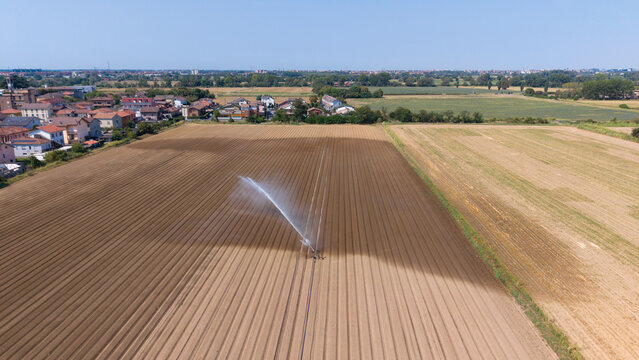 Aerial view of an irrigation sprinkler watering a large brown farmland field with a small town in the background under a clear blue sky in Mediglia, Lombardy, Italy.