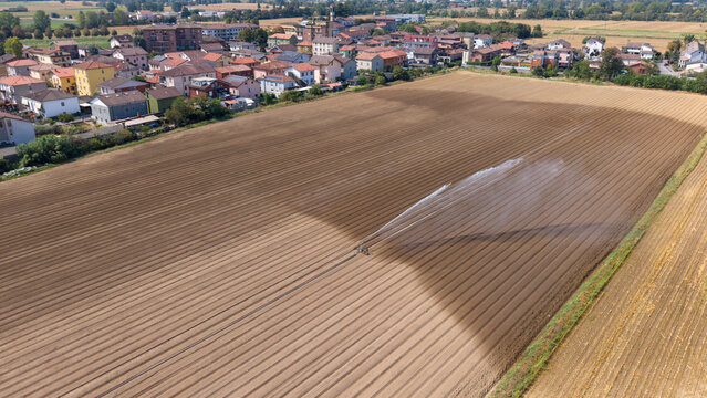 Aerial view of an irrigation sprinkler watering a large plowed field with a small town in the background in Mediglia, Lombardy, Italy.