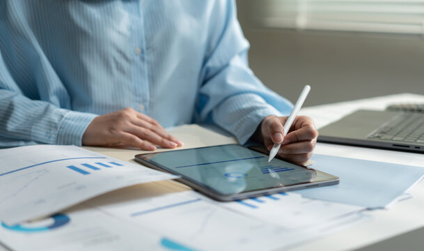 Business analyst using a tablet and stylus with financial reports and laptop, reviewing charts and data for market research, performance analysis, strategic planning and informed decisions.