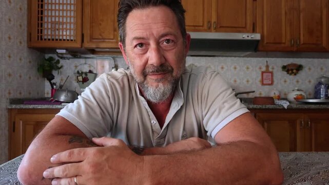 A mature man with a beard sits at a kitchen table, looking at the camera. He slowly transitions from a thoughtful expression to a warm, genuine, and happy smile, conveying joy and contentment in his h