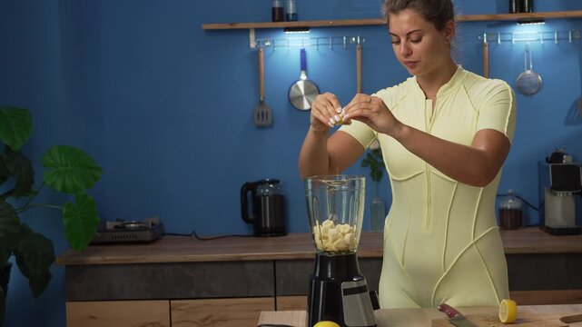 A young, beautiful Caucasian woman in a yellow tracksuit prepares a fruit smoothie in her kitchen at home. She squeezes lemon juice into a blender with the ingredients