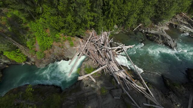 A high overhead aerial view looking straight down at a huge pile of tangled fallen timber logs bridging across churning turquoise river rapids on a bright summer day.