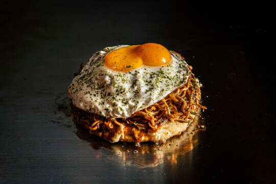 Okonomiyaki pancake with noodles and fried egg on a teppanyaki grill