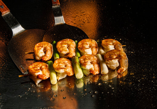 Prawn skewers cooking on a teppanyaki grill with metal spatulas