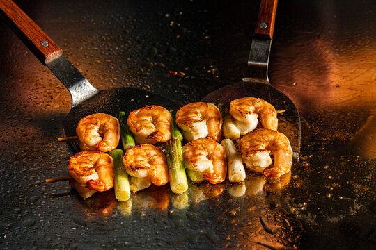 Prawn skewers cooking on a teppanyaki grill with metal spatulas