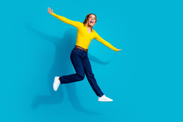 Fototapeta premium Joyful woman jumping against bright blue background wearing yellow sweater and jeans, energetic happy young adult celebrating with arms outstretched