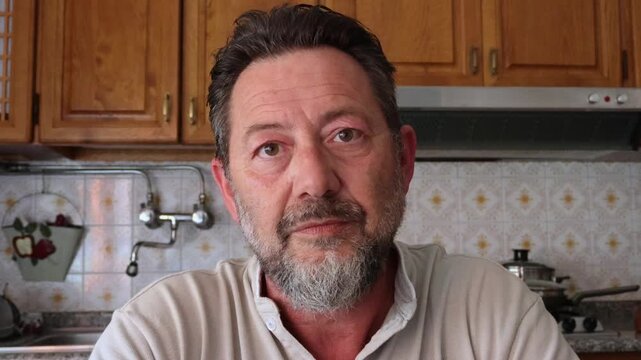 A mature man in a kitchen puts on his eyeglasses, looking directly at the camera. He then gazes thoughtfully around, perhaps focusing on something specific or simply observing his surroundings at home
