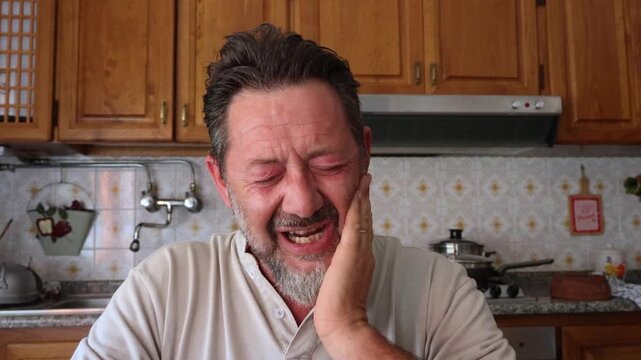 Man in kitchen suffering from terrible toothache, grimacing in pain and holding his cheek. He clearly experiences severe discomfort and needs help for his health problem.