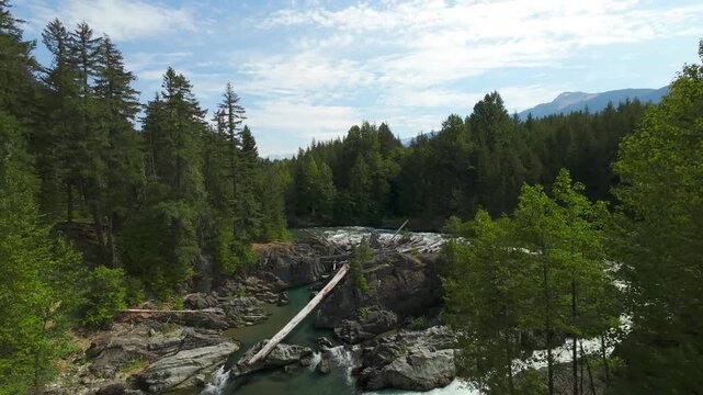 A low aerial camera tracks along a rocky canyon bend, moving over turquoise river pools and white water rapids toward a massive fallen tree log jam on a bright summer day.