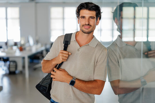 Professional man holding backpack leaning modern office wall