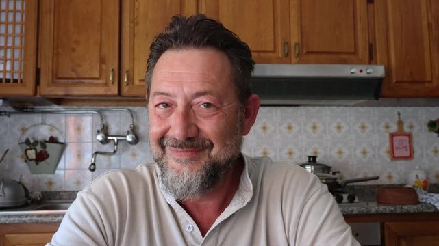 A happy, mature man with a beard smiles warmly at the camera in a cozy kitchen setting, reflecting feelings of contentment and domestic bliss. He looks away and then back, offering genuine expressions