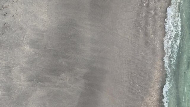 Aerial top down drone view of gentle white wave foam washing onto black sand beach at Nipah Beach, North Lombok, NTB, Indonesia. Crystal clear shallow water meeting dark volcanic sand, abstract natura