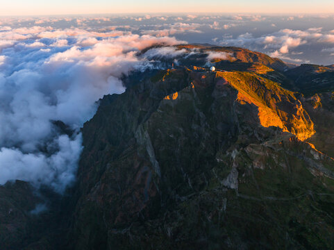 Aerial view of Pico Ruivo mountain peak illuminated by golden hour light above a sea of clouds in Faial, Madeira, Portugal.