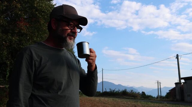 A serene moment captures a bearded man in a cap and glasses enjoying a drink from a metal mug outdoors. He looks thoughtfully at the beautiful mountainous landscape and clear blue sky, embracing the t