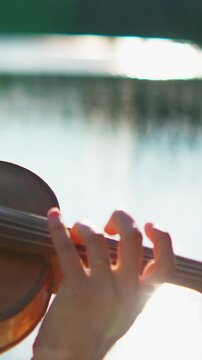 Close up of musician hand gracefully playing violin by tranquil lakeside, fingers pressing strings in warm sunlight creating serene outdoor performance atmosphere