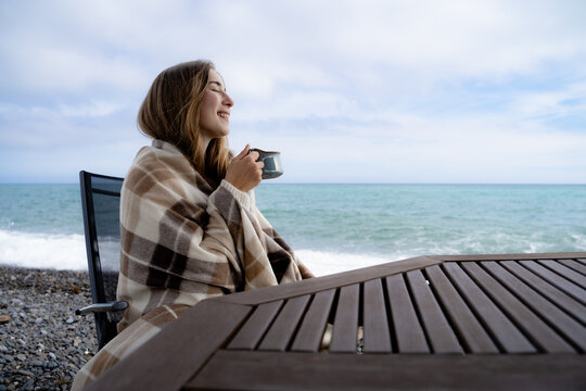 Happy woman wrapped in a cozy blanket, holding a warm beverage and enjoying a peaceful seaside moment on a cloudy day. Leisure time, chilling outdoors in nature.