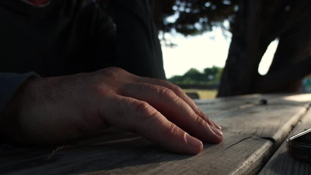 A close-up shot captures a man's hand gently tapping on an old wooden picnic table. Dappled sunlight filters through trees, creating a peaceful and contemplative mood as he rests in nature, perhaps de