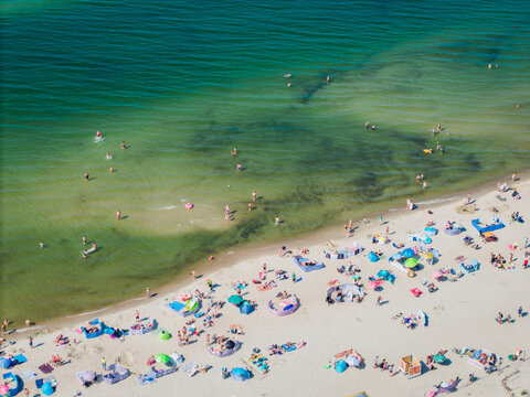 Aerial view of a crowded sandy beach with people swimming in the clear water and relaxing under colorful umbrellas and windbreaks in Krynica Morska, Pomeranian Voivodeshi, Poland.