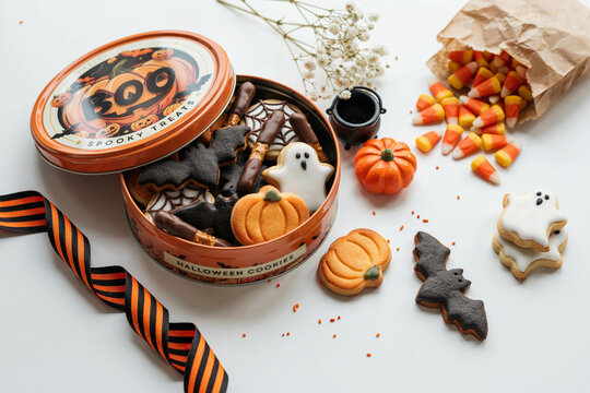 An open orange tin filled with an assortment of spooky Halloween themed cookies shaped like bats ghosts pumpkins and spiderwebs displayed next to decorative candy corn and festive holiday elements