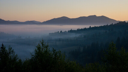 Fototapeta premium Scenic outdoor stock photo of a tranquil morning landscape featuring dense fog rolling over forested hills and distant mountain ridges under a soft dawn sky. Concept for serene nature, eco travel, and
