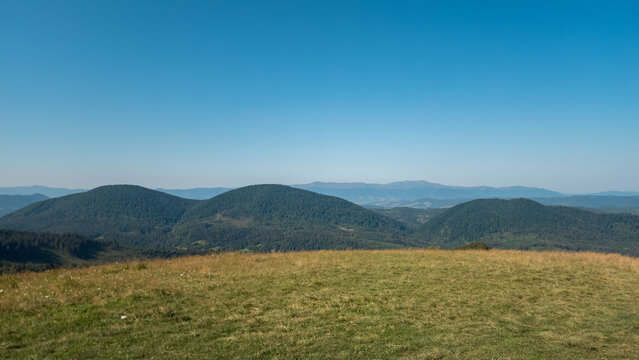 High-angle panoramic view of rolling mountain ridges, sloping grassy hillside, and expansive clear blue sky on a sunny day.