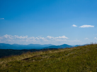 Obraz premium Wide outdoor stock photo of a narrow dirt trail stretching across a grassy alpine mountain ridge toward distant blue peaks under a vast clear blue sky. Concept for trekking journey, eco adventure, tra