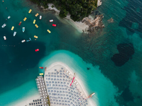 Aerial view of a sandy beach with rows of sun loungers and umbrellas, surrounded by clear turquoise water and small boats in Ksamil, Vlore County, Albania.