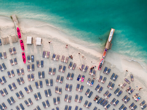Aerial view of a sandy beach lined with rows of sun loungers and colorful kayaks resting on a wooden pier, surrounded by clear turquoise water in Ksamil, Vlore County, Albania.
